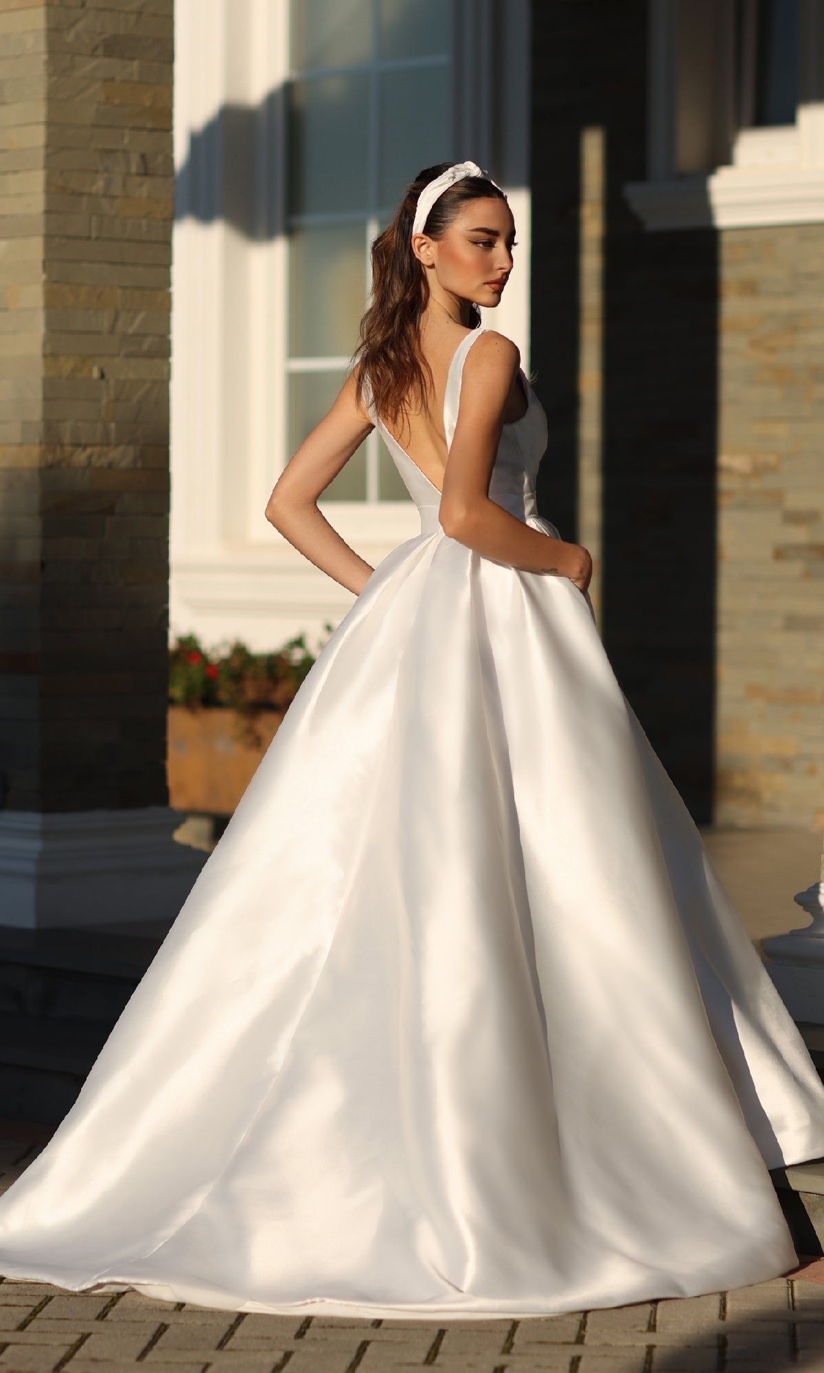 A woman wearing the Tina Holly BB209W belted long white prom dress stands outdoors near a stone-walled building, looking over her shoulder.